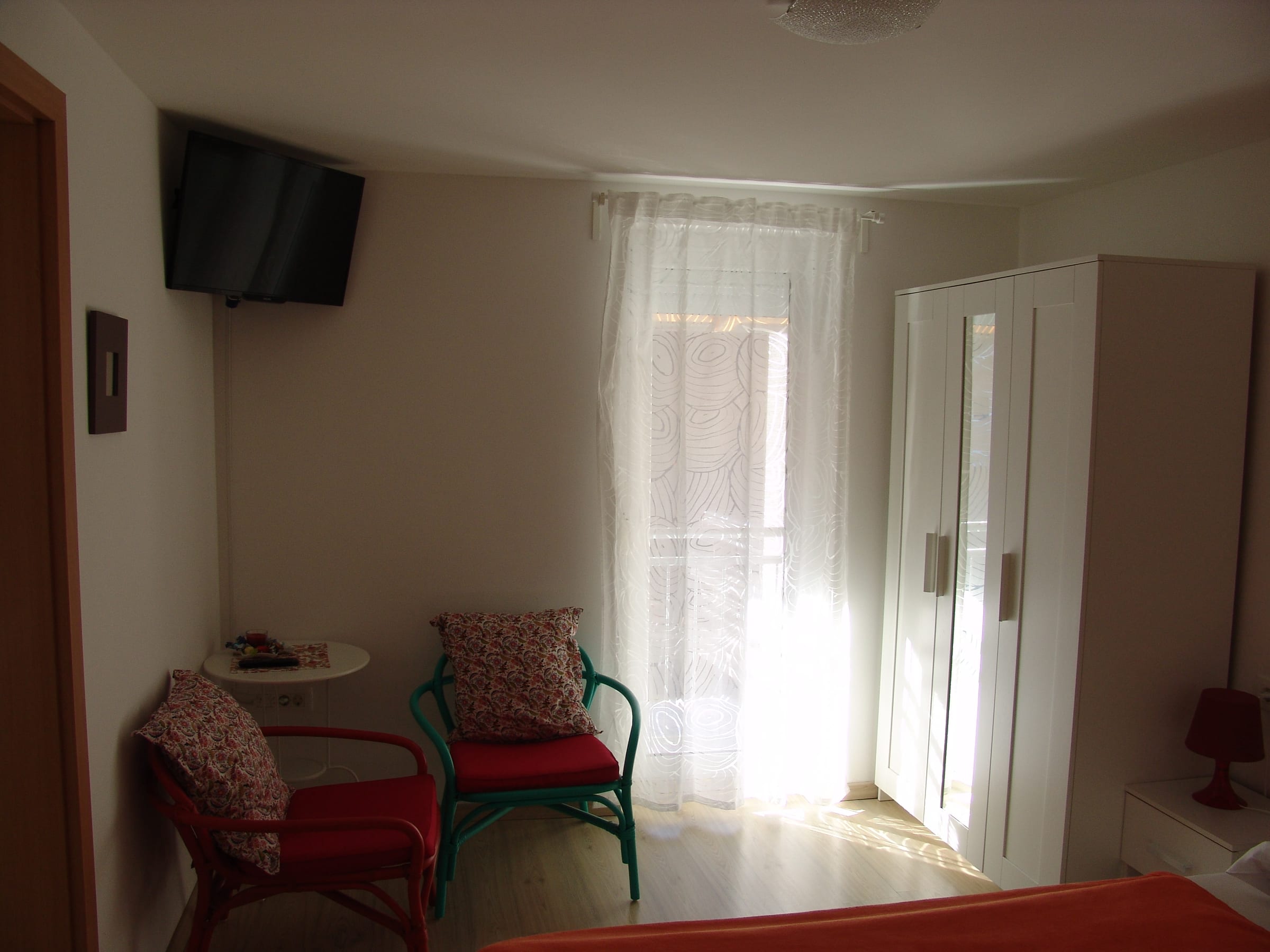 Two armchairs and a small table by a curtained window, with a wall-mounted television and a white wardrobe.