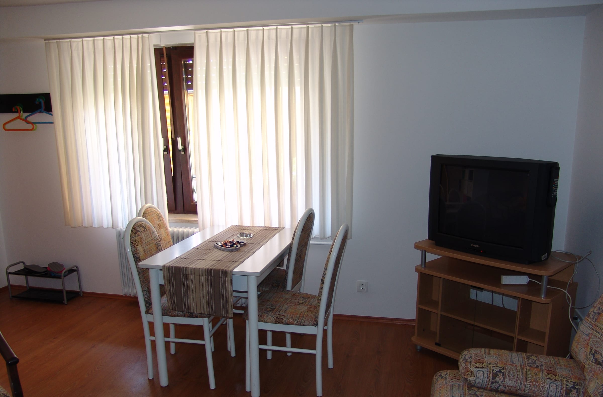 A living and dining area with a table and chairs, a television on a wooden stand and a bright curtained window.